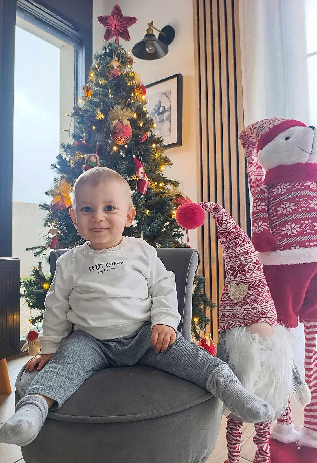 Ayden participe au concours pour gagner de l'argent avec cette photo : child, toddler, baby, christmas_tree, ornament, lights, chair, armchair, gnome, plush, festive, holiday, smile, sweater, socks, living_room, decoration, portrait, happy, indoor