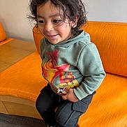Izayah is registered to the contest to win money with this photo: child, curly_hair, green_hoodie, toy_car, orange_couch, black_pants, sneakers, smiling, indoor, seated, happy, casual_clothing, person, kid, playful, cute, young, portrait, cozy, relaxed