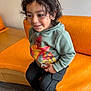 child, curly_hair, green_hoodie, toy_car, orange_couch, black_pants, sneakers, smiling, indoor, seated, happy, casual_clothing, person, kid, playful, cute, young, portrait, cozy, relaxed