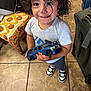 Izayah is registered to the contest to win money with this photo: child, curly_hair, smiling, toy_car, indoor, tiled_floor, shirt, sneakers, person, happy, standing, playful, shorts, luggage, chair, blanket, floor, holding, cute, young