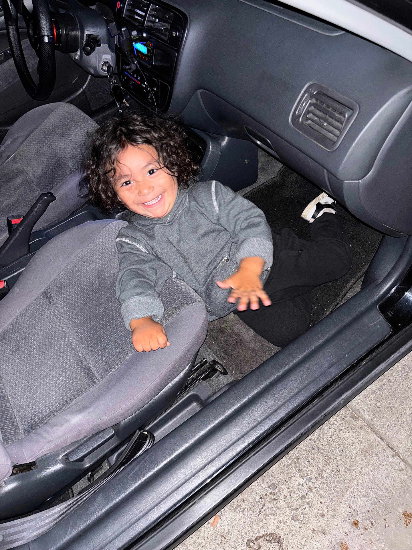 Izayah joined the competition — help win amazing prizes! child, smiling, curly_hair, car_interior, car_seat, floor, grey_clothing, black_pants, shoe, hand, dashboard, seatbelt, steering_wheel, car_door, concrete, playful, happy, person, indoor, vehicle