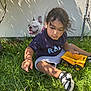 child, grass, toy_truck, dog, french_bulldog, outdoor, sunlight, shadow, wall, greenery, playing, casual_clothing, shorts, tshirt, shoe, pet, summer, nature, happy_dog, curly_hair