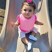 Aria joined the competition — help win amazing prizes! toddler, child, slide, playground, pink_bow, pink_bib, curly_hair, white_shoes, sunlight, shadow, outdoor, play, cute, baby, sitting, happy, park, clothing, person, smile