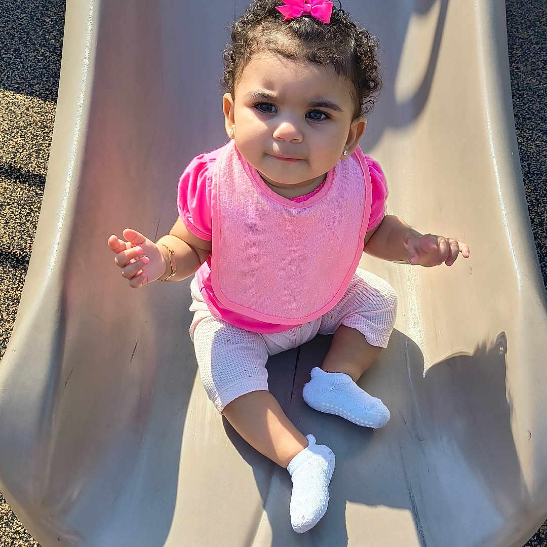 Aria joined the competition — help win amazing prizes! toddler, child, slide, playground, pink_bow, pink_bib, curly_hair, white_shoes, sunlight, shadow, outdoor, play, cute, baby, sitting, happy, park, clothing, person, smile