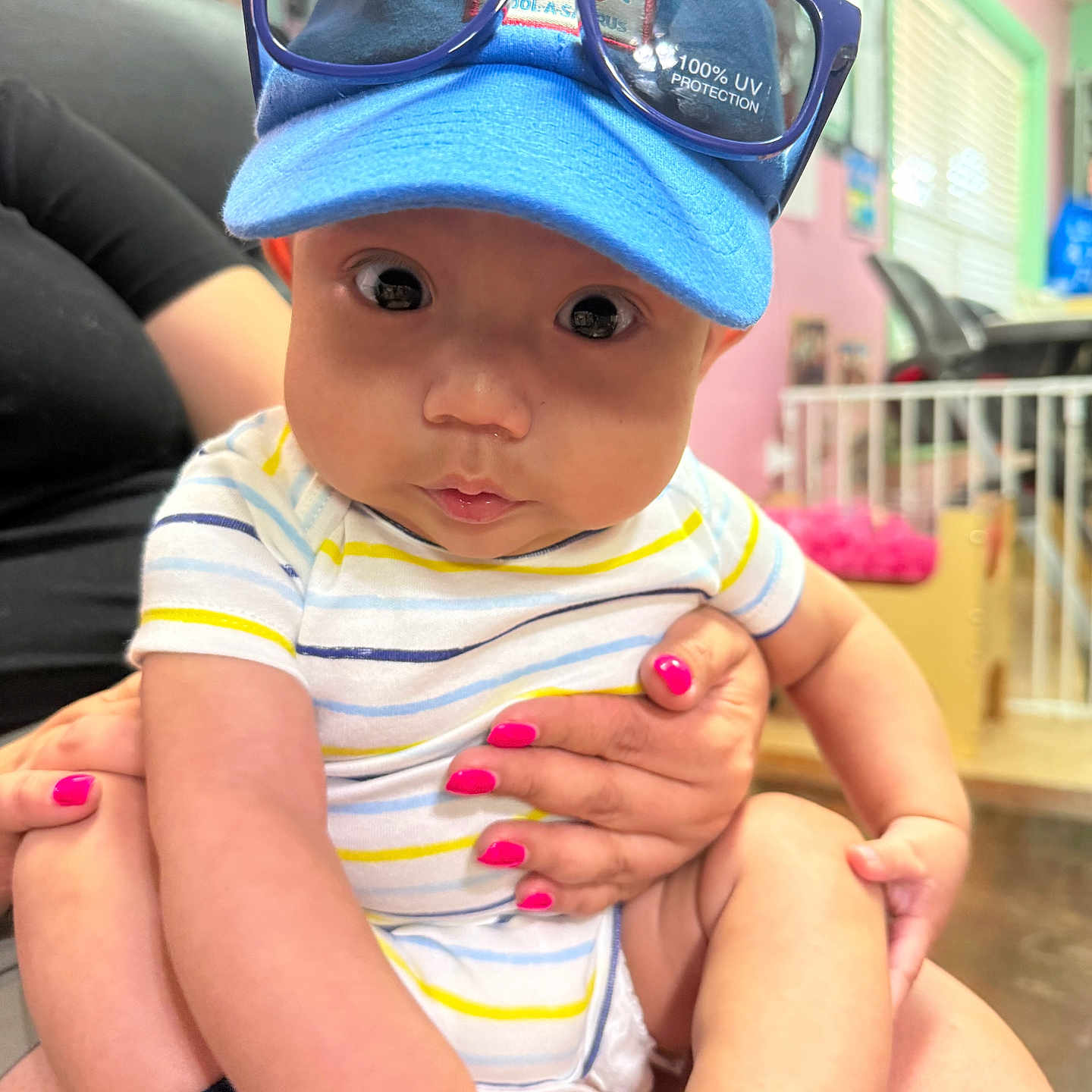Samuel is registered to the contest to win money with this photo: baby, infant, child, blue_cap, sunglasses, striped_onesie, pink_nails, hand, indoor, person, curious, cute, seated, holding, furniture, colorful, bright, face, sock, expression