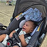 baby, car_seat, child, infant, smiling, happy, outdoor, person, seatbelt, clothing, fabric, blanket, tree, fence, pavement, sunlight, relaxation, park, casual, seated