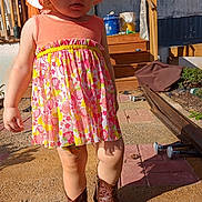Hazel is registered to the contest to win money with this photo: child, toddler, dress, hat, cowboy_boots, outdoor, sunlight, garden, wooden_structure, concrete, pavement, person, footwear, clothing, summer, daylight, expression, standing, colorful, young