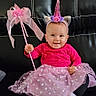 baby, child, unicorn_costume, headband, tutu, pink, polka_dots, wand, smiling, sitting, leather_couch, indoor, cute, happy, costume, dress, footwear, black_background, portrait, person