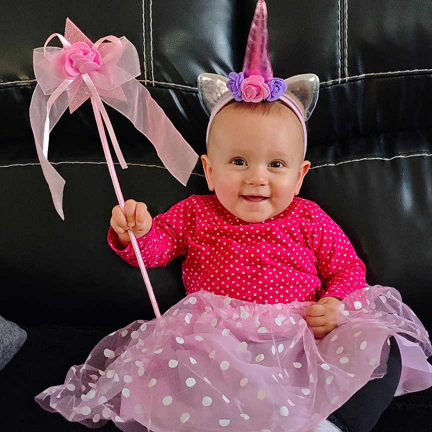 Ophélya participe au concours pour gagner de l'argent avec cette photo : baby, child, unicorn_costume, headband, tutu, pink, polka_dots, wand, smiling, sitting, leather_couch, indoor, cute, happy, costume, dress, footwear, black_background, portrait, person