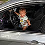 toddler, child, car, steering_wheel, diaper, tshirt, smiling, inside_car, driver_seat, pointing, silver_car, happy, young_child, automobile_interior, seat, window, door_handle, outdoor, greenery, playful