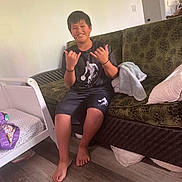 Kymani joined the competition — help win amazing prizes! barefoot, basketball_graphic, blanket, boy, casual_clothing, child, couch, crib, flooring, green_couch, hand_gesture, happy, indoor, living_room, pillow, shaka_sign, shorts, smile, tshirt, wood_floor