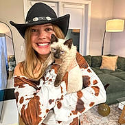 Cowboy Charlie joined the competition — help win amazing prizes! woman, cat, siamese_cat, cowboy_hat, smiling, indoor, living_room, lamp, couch, pillow, rug, table, balloon, mirror, blonde_hair, long_hair, shirt, holding, pet, happy