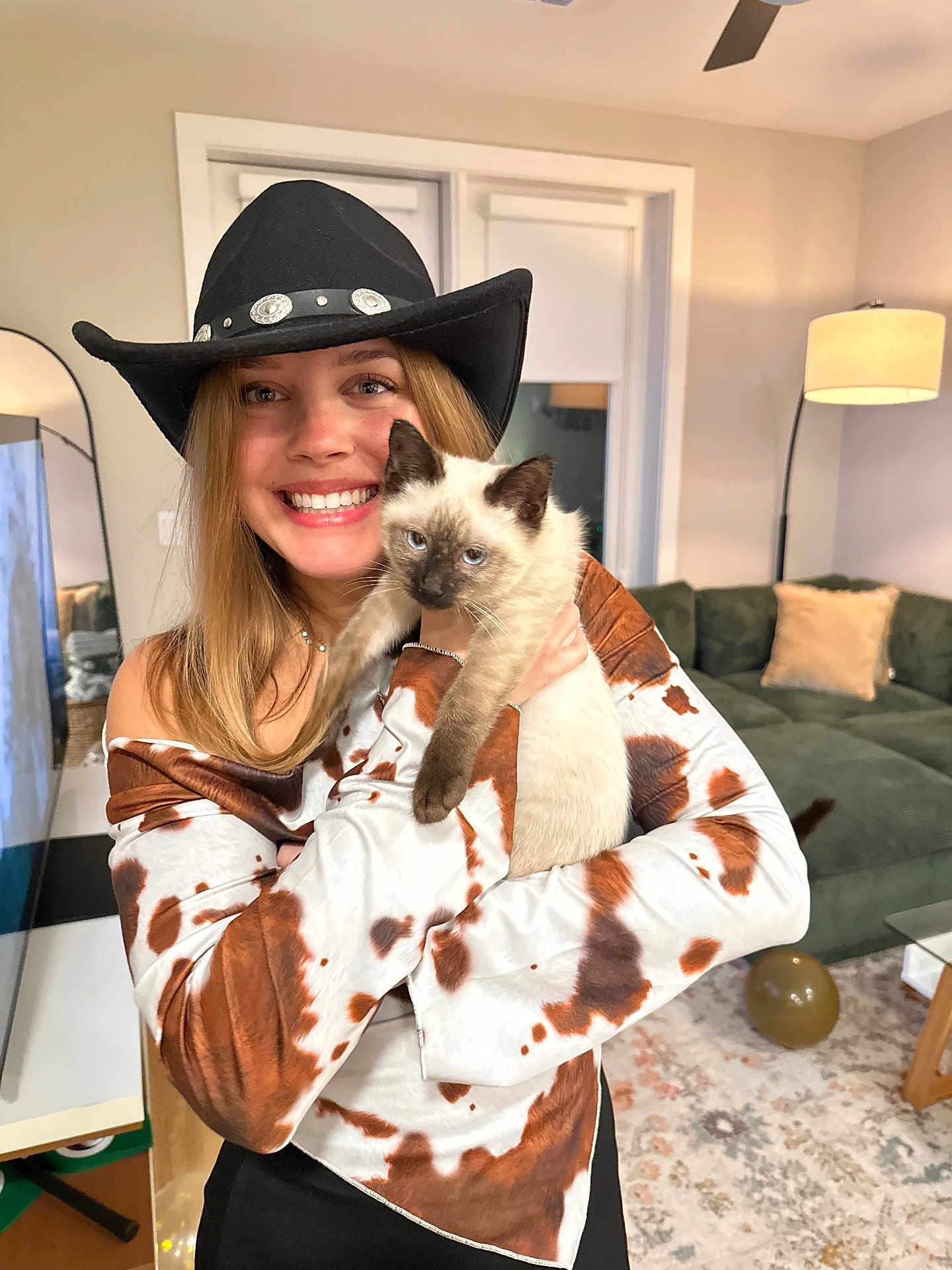 Cowboy Charlie joined the competition — help win amazing prizes! woman, cat, siamese_cat, cowboy_hat, smiling, indoor, living_room, lamp, couch, pillow, rug, table, balloon, mirror, blonde_hair, long_hair, shirt, holding, pet, happy