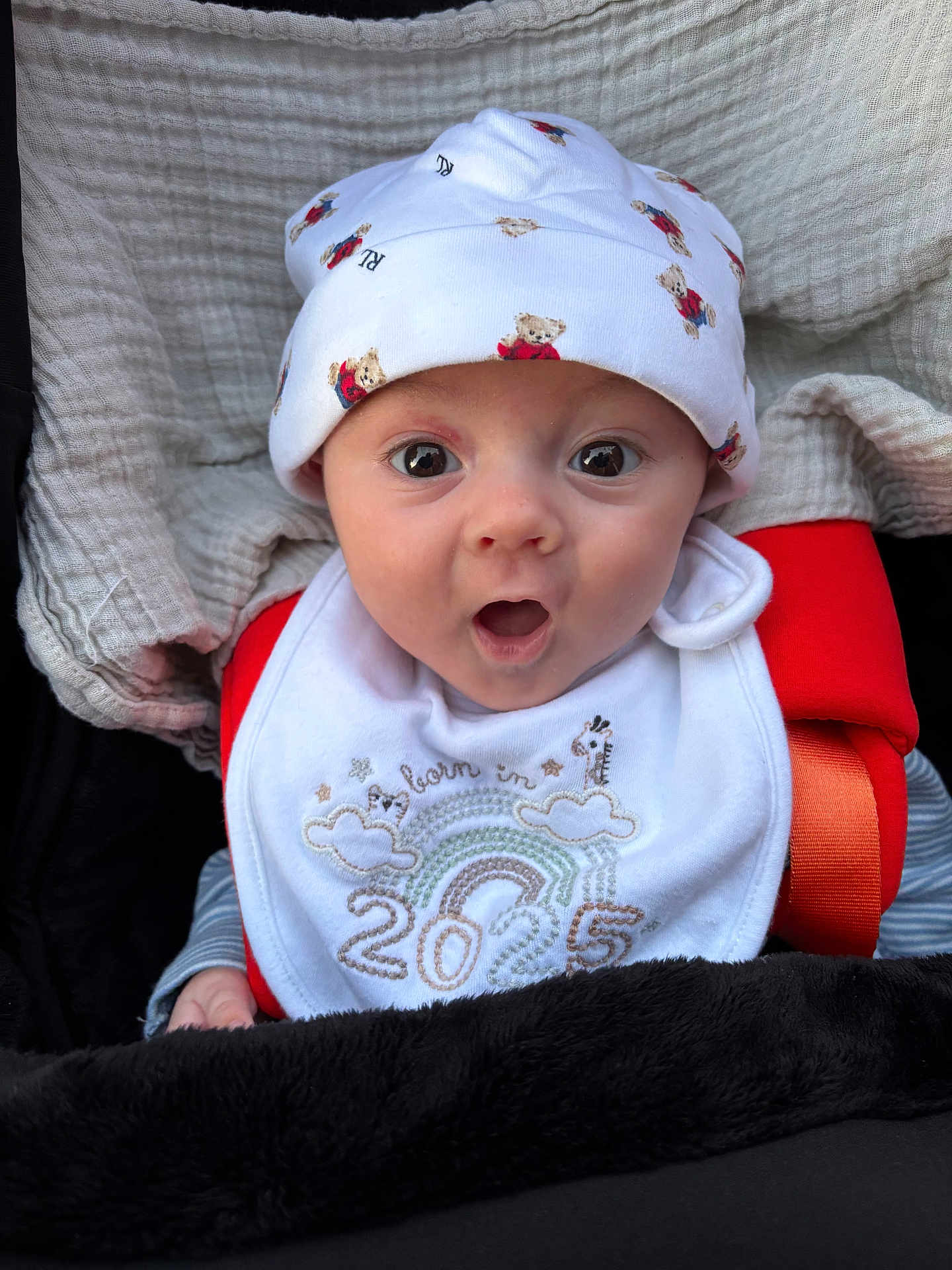 Kai is registered to the contest to win money with this photo: baby, infant, hat, bib, blanket, clothing, face, wide_eyes, expression, cute, teddy_bear_pattern, newborn, portrait, child, sitting, cozy, soft_texture, red_strap, mouth_open, cute_expression