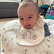 Kai joined the competition — help win amazing prizes! baby, smile, bib, cushion, blanket, indoor, toy, window, natural_light, patterned_clothing, child, infant, happy, face, soft_texture, home, colorful, cute, playtime, cozy