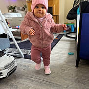 Milena is registered to the contest to win money with this photo: toddler, child, pink_jacket, pink_hat, sneakers, indoor, living_room, toy_car, rug, flooring, furniture, fireplace, baby_toys, baby, person, walking, cute, cozy, home, portrait