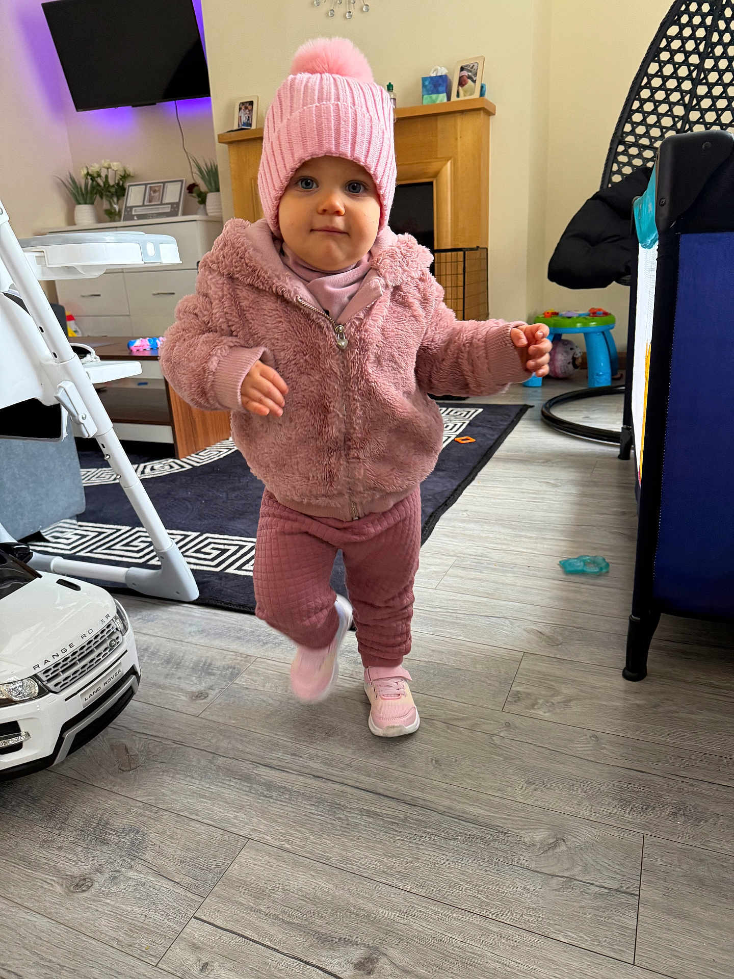 Milena is registered to the contest to win money with this photo: toddler, child, pink_jacket, pink_hat, sneakers, indoor, living_room, toy_car, rug, flooring, furniture, fireplace, baby_toys, baby, person, walking, cute, cozy, home, portrait