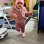 toddler, child, pink_hat, pink_jacket, pink_pants, pink_shoes, indoor, living_room, toy_car, television, wooden_floor, rug, furniture, baby, person, walking, cute, warm_clothing, home, floor