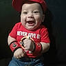 apparel, baby, child, clothing, cute, denim_shorts, face, happy, indoor, infant, motivation, person, portrait, positive_message, red_cap, red_shirt, sitting, smiling, wristbands, young_child