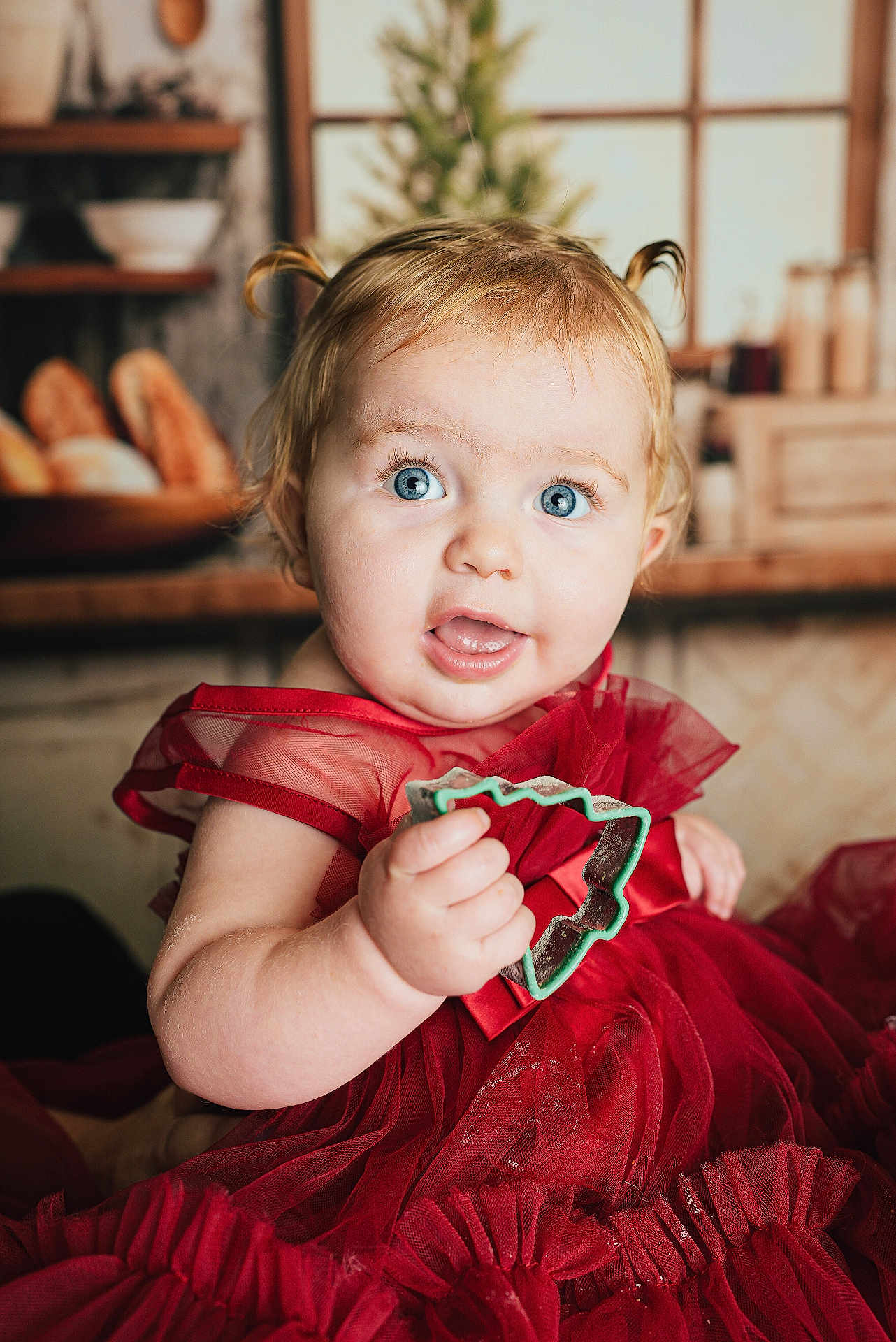 Elodie joined the competition — help win amazing prizes! baby, child, blue_eyes, red_dress, cookie_cutter, kitchen, indoor, portrait, cute, toddler, curly_hair, holding, expression, face, person, smile, furniture, background, window, sitting