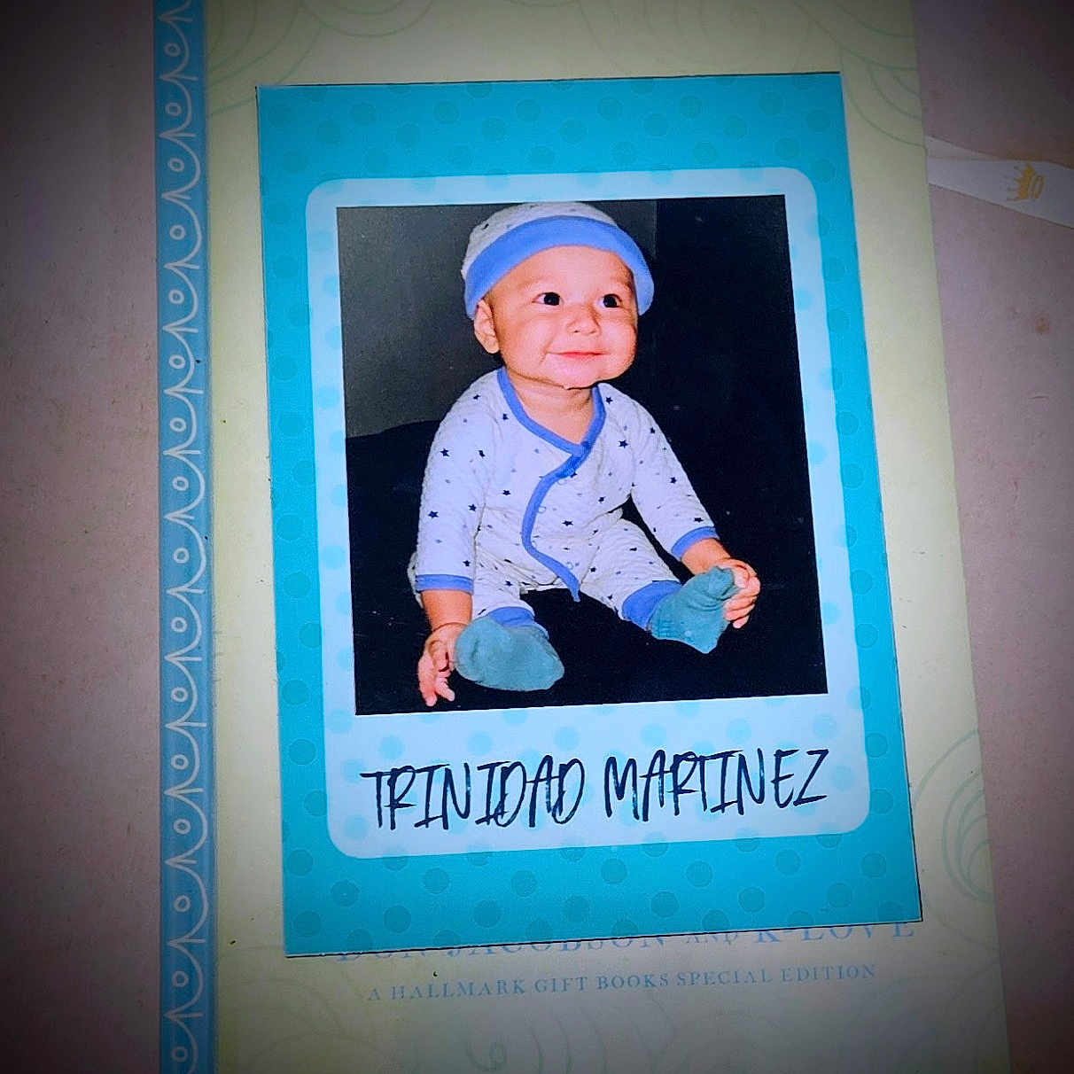 Trinidad is registered to the contest to win money with this photo: baby, child, infant, portrait, smiling, happy, sitting, blue_hat, onesie, stars, socks, printed_photo, keepsake, scrapbook, book, paper, blue_frame, indoor, closeup, family_memento