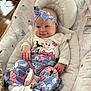 baby, infant, smiling, headband, clothing, pants, socks, baby_seat, floral_pattern, indoor, blanket, couch, wood_floor, cute, comfort, person, face, hand, footwear, head