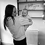 Ruby is registered to the contest to win money with this photo: woman, baby, smile, indoor, black_and_white, holding, joy, person, sweatshirt, leggings, radiator, shelf, slippers, floor_tile, wall_art, happy, face, hair, home, portrait