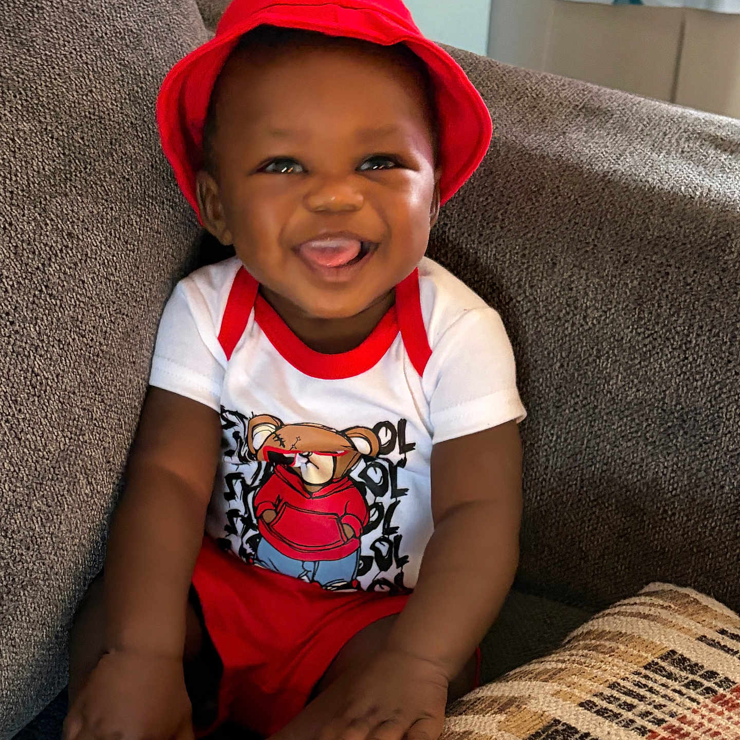 Jarriett joined the competition — help win amazing prizes! baby, child, smiling, red_hat, red_shorts, white_shirt, cartoon_tshirt, sitting, couch, gray_couch, plush, happy, tongue_out, indoors, cute, portrait, young_child, infant, homedecor, cozy