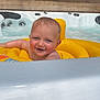 baby, child, smiling, water, inflatable_ring, hot_tub, pool, outdoor, sunlight, wet_hair, yellow, joyful, face, floating, summer, playful, bathing, relaxation, water_bubbles, deck