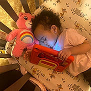 Zaniyah-Ann joined the competition — help win amazing prizes! toddler, child, crib, tablet, plush_toy, care_bear, winnie_the_pooh, bed, sleeping, earrings, pink_clothing, patterned_bedding, toy, indoor, cozy, person, cute, curly_hair, resting, technology
