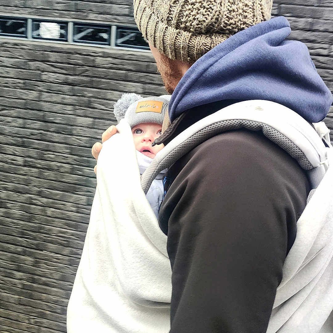Hortense a rejoint le concours — aidez-le/la à gagner de superbes lots ! baby, adult, hat, knitted_hat, pom_pom, blanket, baby_carrier, outdoor, brick_wall, person, face, clothing, cold_weather, hoodie, jacket, carrying, walking, child, winter, cozy