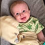baby, smiling, infant, child, green_clothing, blanket, toy, chair, happy, cute, portrait, indoors, soft_furniture, palm_tree_pattern, holding, waving, playful, young_child, comfort, bright_eyes