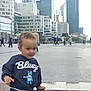 toddler, child, bluey, sweatshirt, granite, ledge, urban, city, skyscrapers, pedestrians, people, sidewalk, curly_hair, casual_clothing, outdoor, daytime, playful, exploration, young_child, portrait