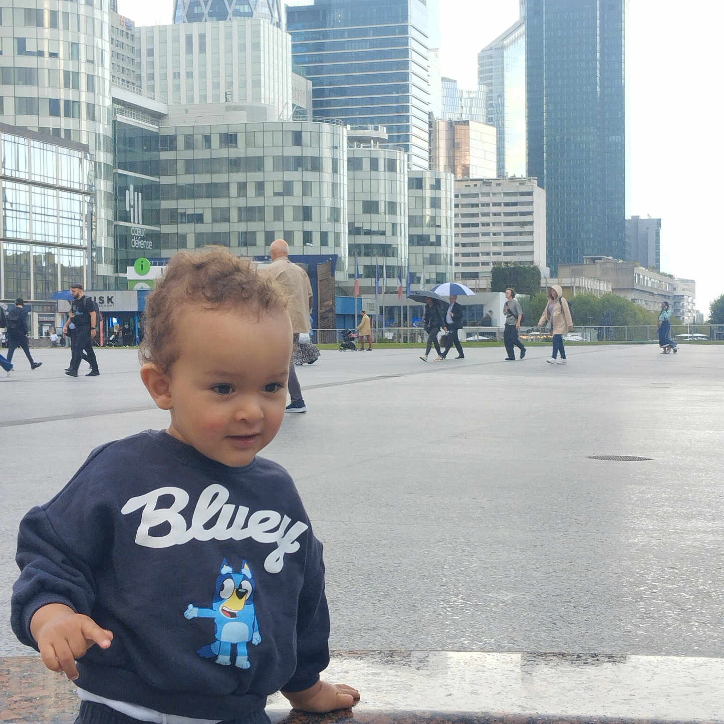 Ayden a rejoint le concours — aidez-le/la à gagner de superbes lots ! bluey, casual_clothing, child, city, curly_hair, daytime, exploration, granite, ledge, outdoor, pedestrians, people, playful, portrait, sidewalk, skyscrapers, sweatshirt, toddler, urban, young_child