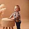 toddler, child, smile, standing, stool, woven_stool, plaid_shirt, jeans, socks, beige_background, studio, happy, cute, indoor, person, furniture, decor, fluffy_plant, portrait, casual_clothing