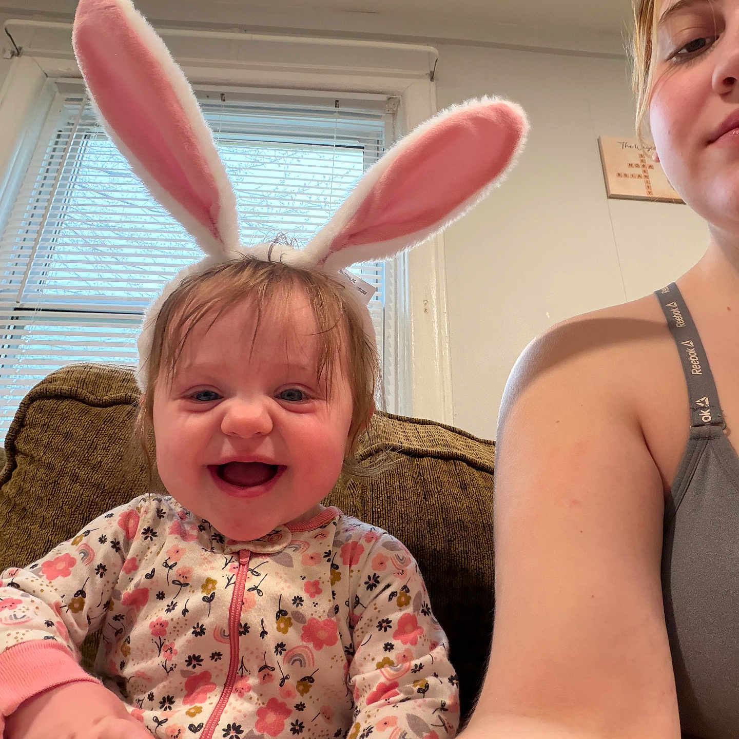 Nora is registered to the contest to win money with this photo: baby, bunny_ears, smiling, woman, couch, window, indoor, child, happy, pink, face, person, blinds, casual_clothing, home, portrait, sitting, cute, young_child, fun