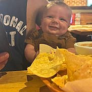 Ryker is registered to the contest to win money with this photo: baby, smile, person, adult, tortilla_chips, food, table, basket, dip, salsa, restaurant, indoor, wooden_table, clothing, sleeveless_shirt, face, hand, happy, casual, dining