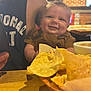 baby, smile, person, adult, tortilla_chips, food, table, basket, dip, salsa, restaurant, indoor, wooden_table, clothing, sleeveless_shirt, face, hand, happy, casual, dining
