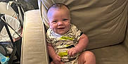 Ryker joined the competition — help win amazing prizes! baby, child, smiling, couch, onesie, dinosaur_print, furniture, indoor, happy, face, person, infant, seated, plush, home, casual, armrest, leg, foot, skin