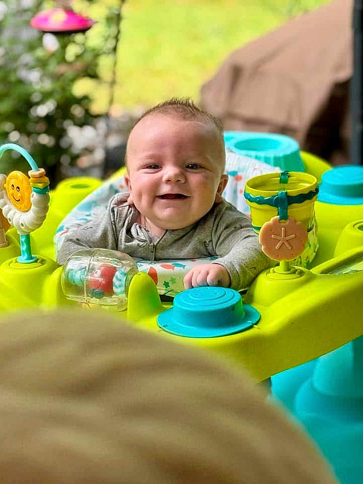 Ryker is registered to the contest to win money with this photo: baby, smiling, activity_center, toy, colorful, infant, face, happy, child, play, greenery, outdoor, blurred_background, cute, young_child, portrait, seat, hands, clothing, indoor