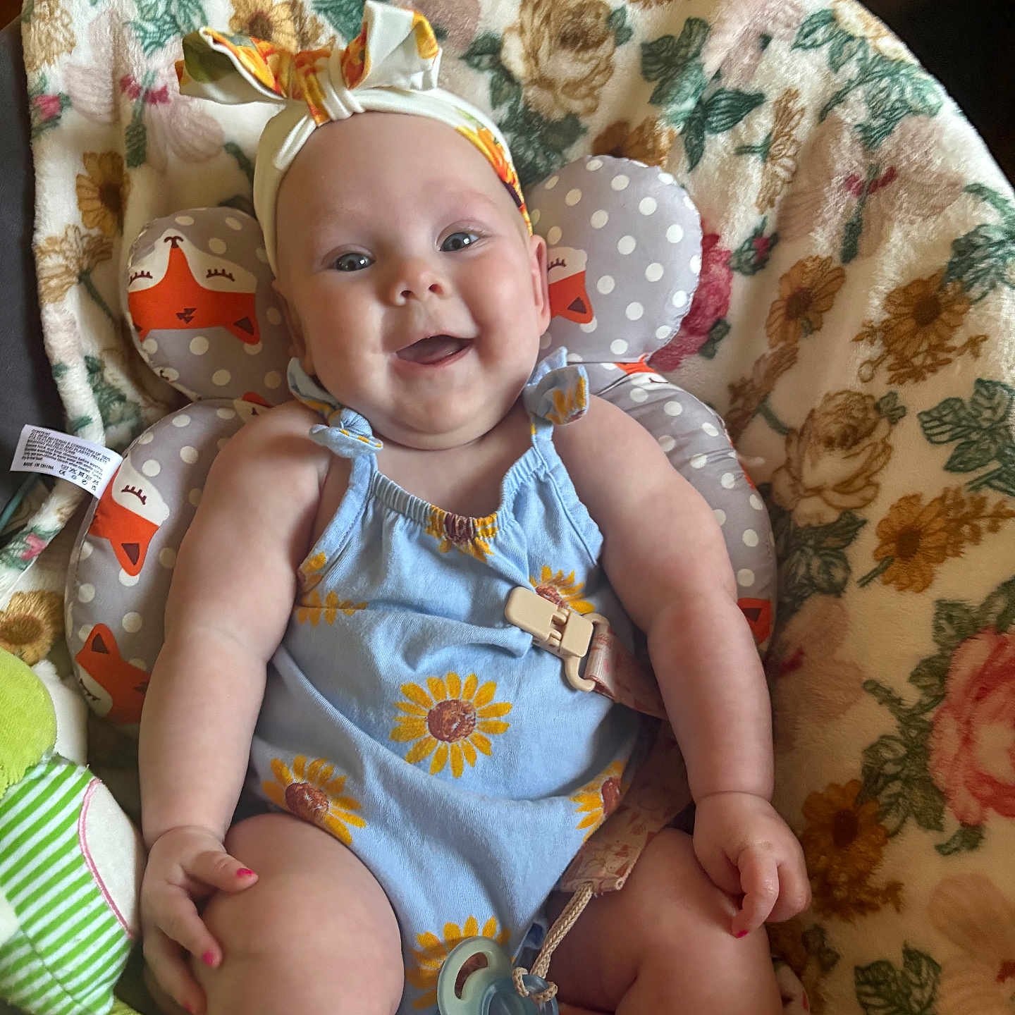 Berkley is registered to the contest to win money with this photo: baby, child, smiling, romper, headband, bow, flower_pattern, sunflower, cushion, fox_pattern, blanket, floral, sitting, indoors, cute, happy, infant, person, face, pacifier