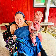 Berkley is registered to the contest to win money with this photo: accessories, baby, bench, blue_dress, cute, hand, happy, headband, holding, infant, outdoor, pacifier, person, red_wall, seated, smile, tattoo, wheelchair, window, woman