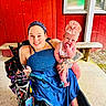 accessories, baby, bench, blue_dress, cute, hand, happy, headband, holding, infant, outdoor, pacifier, person, red_wall, seated, smile, tattoo, wheelchair, window, woman
