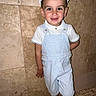 child, boy, smile, overalls, white_shirt, cap, standing, tile_wall, indoor, happy, portrait, casual_clothing, shorts, fashion, cute, person, young, playful, light_blue, background