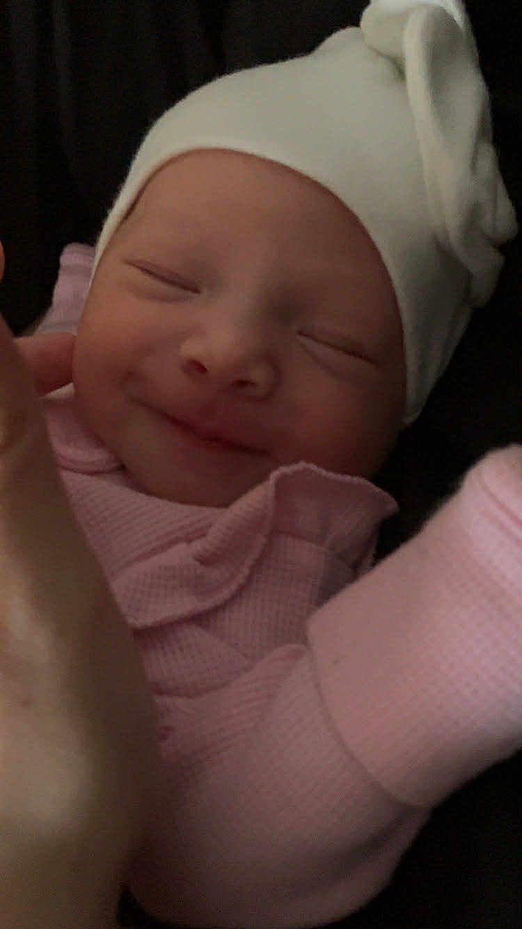 Amelia is registered to the contest to win money with this photo: baby, newborn, smiling, sleepy, hat, flower, pink_clothing, pajamas, hand, closeup, face, infant, cute, cozy, resting, child, portrait, soft_light, peaceful, indoors