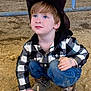 child, boy, cowboy_hat, blue_eyes, plaid_shirt, jeans, boots, dirt, arena, crouching, outdoor, curious, casual_clothing, rustic, young, portrait, hat, hands, expression, small_stick