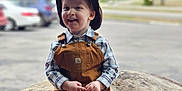 Brantley is registered to the contest to win money with this photo: toddler, child, boy, cowboy_hat, overalls, plaid_shirt, smiling, sitting, rock, outdoor, parking_lot, cars, sign, daylight, happy, casual_clothing, boots, portrait, young_child, playful