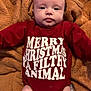 baby, infant, onesie, red_clothing, text_on_clothing, blanket, brown_blanket, lying_down, face, legs, arms, cute, child, portrait, indoors, soft_texture, holiday_theme, christmas, expression, newborn