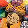 Jaxson is registered to the contest to win money with this photo: baby, infant, child, three_months, sign, wooden_sign, yellow_clothing, cushions, patterned_cushions, soft_toys, indoor, portrait, smiling, hands_clasped, seated, face, head, cozy, colorful, cute