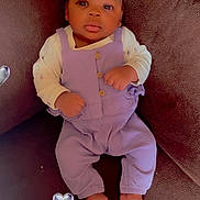 Kanyla is registered to the contest to win money with this photo: baby, infant, purple_overalls, cream_shirt, brown_couch, face, barefoot, heart_sticker, indoor, child, cute, portrait, looking_at_camera, relaxed, seated, hands_clenched, dark_skin, short_hair, soft_lighting, cozy